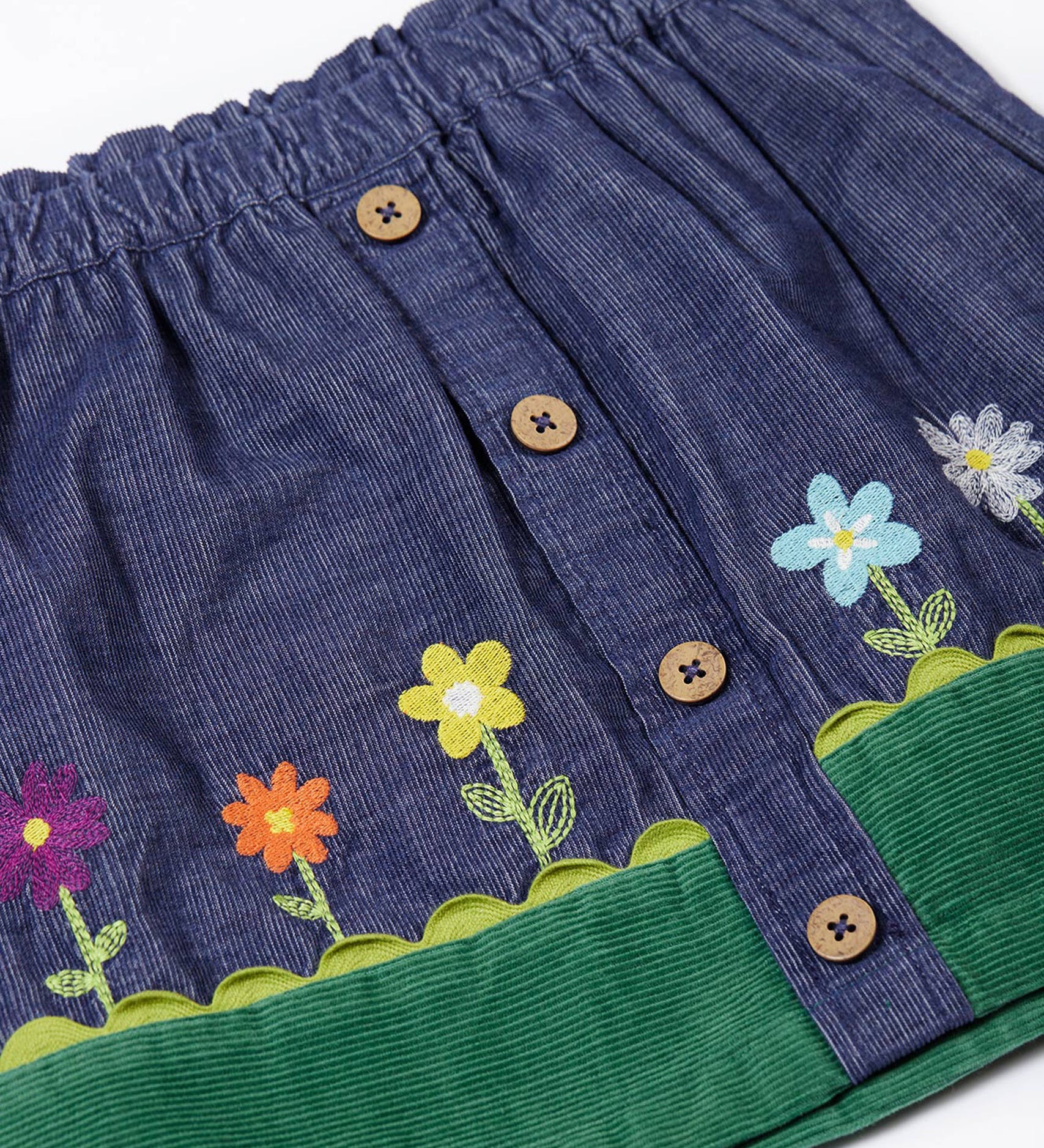 A close up of the front of the Frugi Cara cord children's skirt in ink blue with flower and grass details. The skirt has a green band with a scalloped edge to create the look of grass,