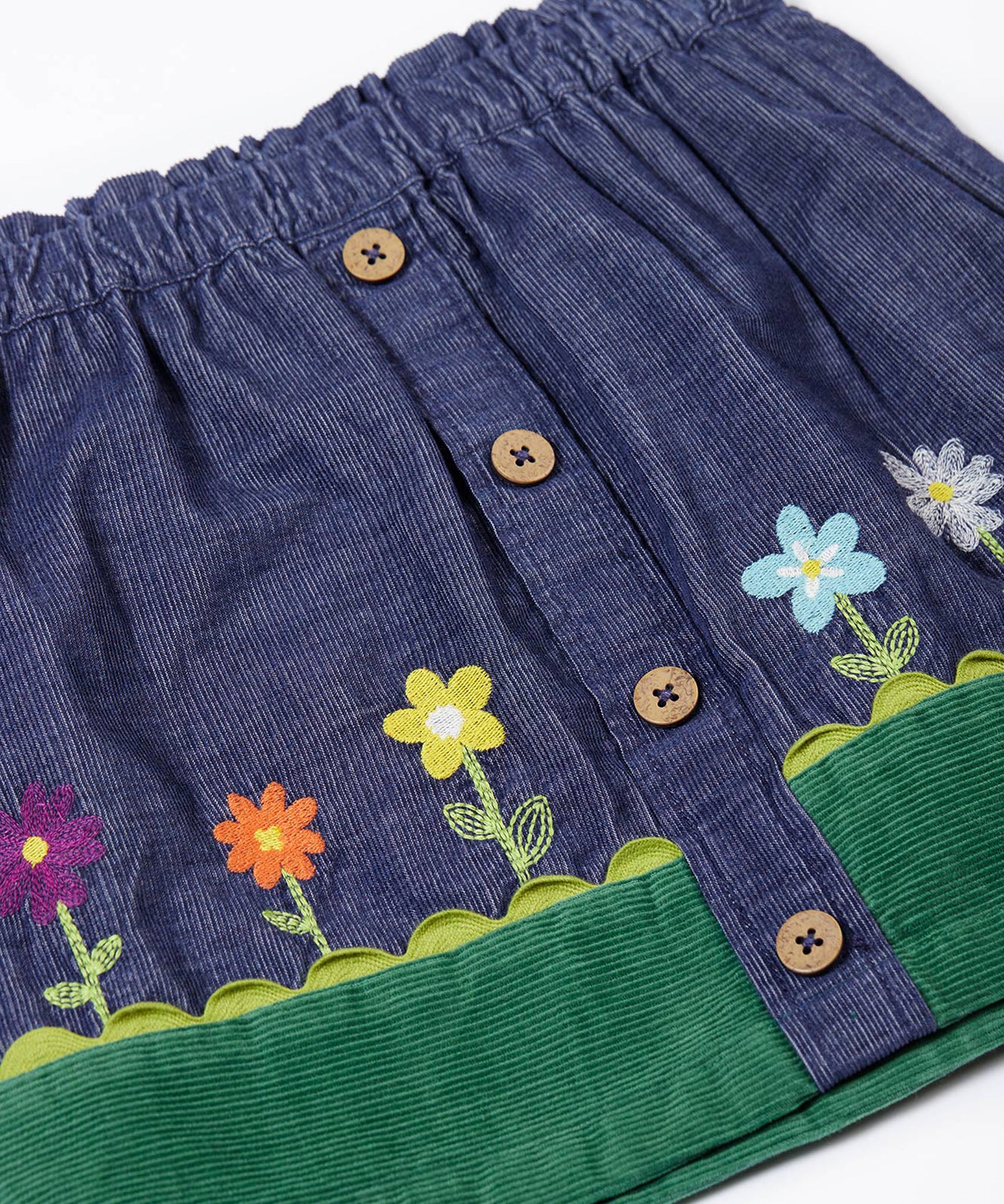 A close up of the front of the Frugi Cara cord children's skirt in ink blue with flower and grass details. The skirt has a green band with a scalloped edge to create the look of grass,