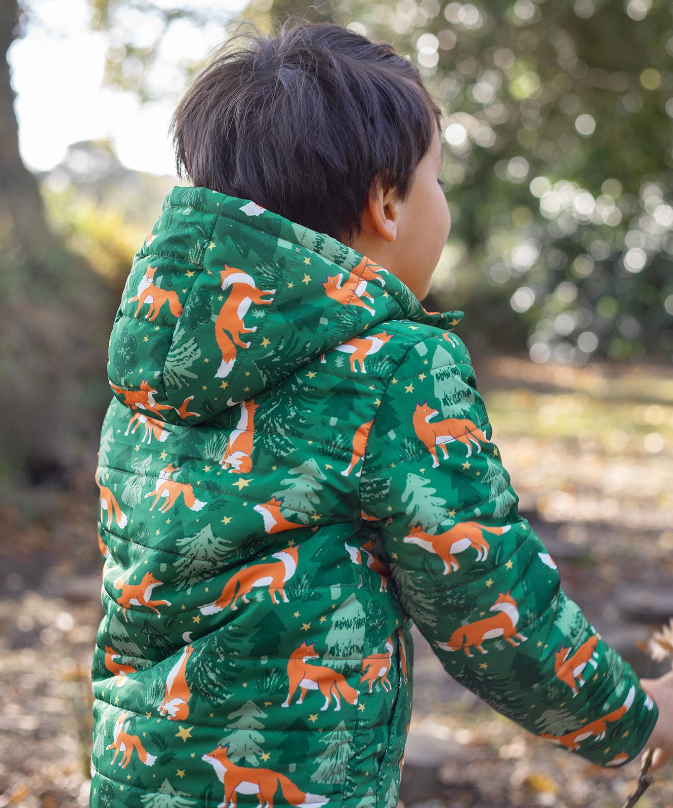 A child wearing the Frugi toasty trail reversible jacket showing the playful fox in woodland design and hood detail on green post consumer recycled polyester fabric