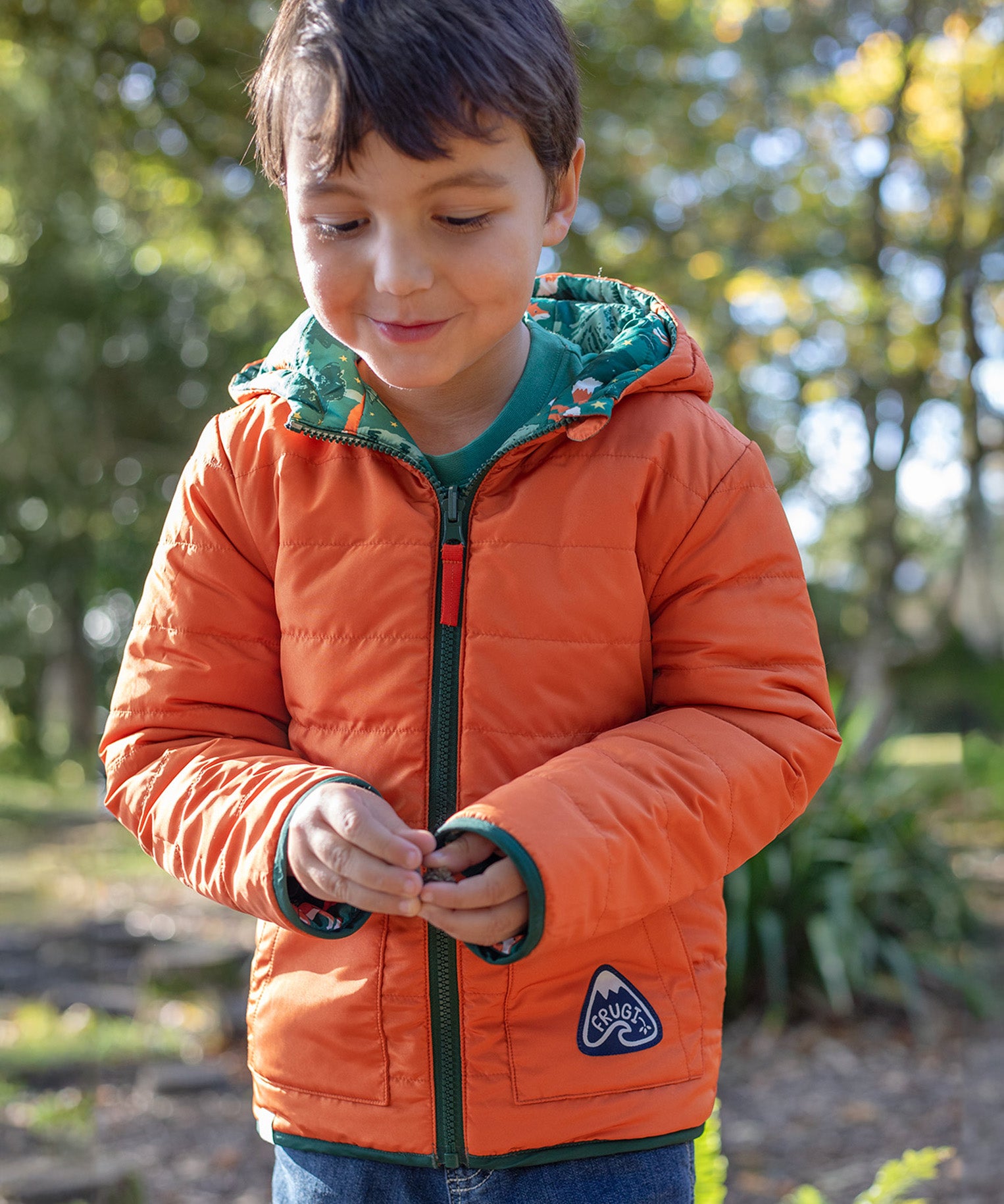 A child wearing the Frugi toasty trail reversible jacket showing the orange colour side and green zip detail with a brand patch on the side pocket on post consumer recycled polyester fabric