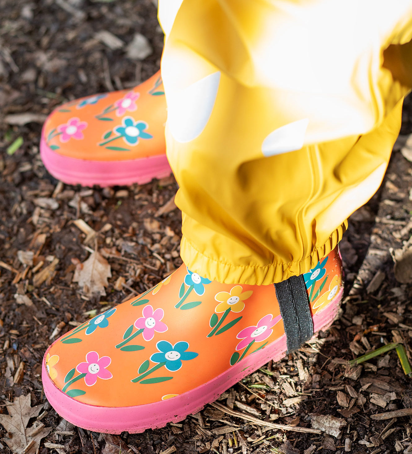 A child standing outside and wearing the Frugi puddle buster yellow trousers and puddle buster wellington boots in fun flowers  A natural rubber and waterproof wellington boot in orange with prints of multicoloured, playful flowers.