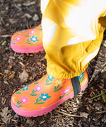 A child standing outside and wearing the Frugi puddle buster yellow trousers and puddle buster wellington boots in fun flowers  A natural rubber and waterproof wellington boot in orange with prints of multicoloured, playful flowers.