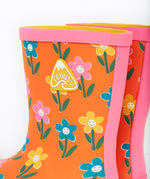 A close up of the top of the Frugi kids puddle buster wellington boots. Showing the the pink trim and flower print detail on orange natural rubber material. Features a yellow triangular brand patch on the outside of the boot. 