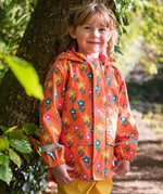 A toddler standing by a tree and wearing the Frugi puddle buster yellow trousers, fun flowers coat.  An orange waterproof coat with playful prints of flowers.