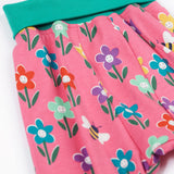A close up of the print of the Frugi fun flowers parsnip pants. The fun flowers print features multi-coloured flowers and bees on a pink background with turquoise coloured waistband and leg cuffs.