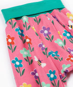 A close up of the print of the Frugi fun flowers parsnip pants. The fun flowers print features multi-coloured flowers and bees on a pink background with turquoise coloured waistband and leg cuffs.