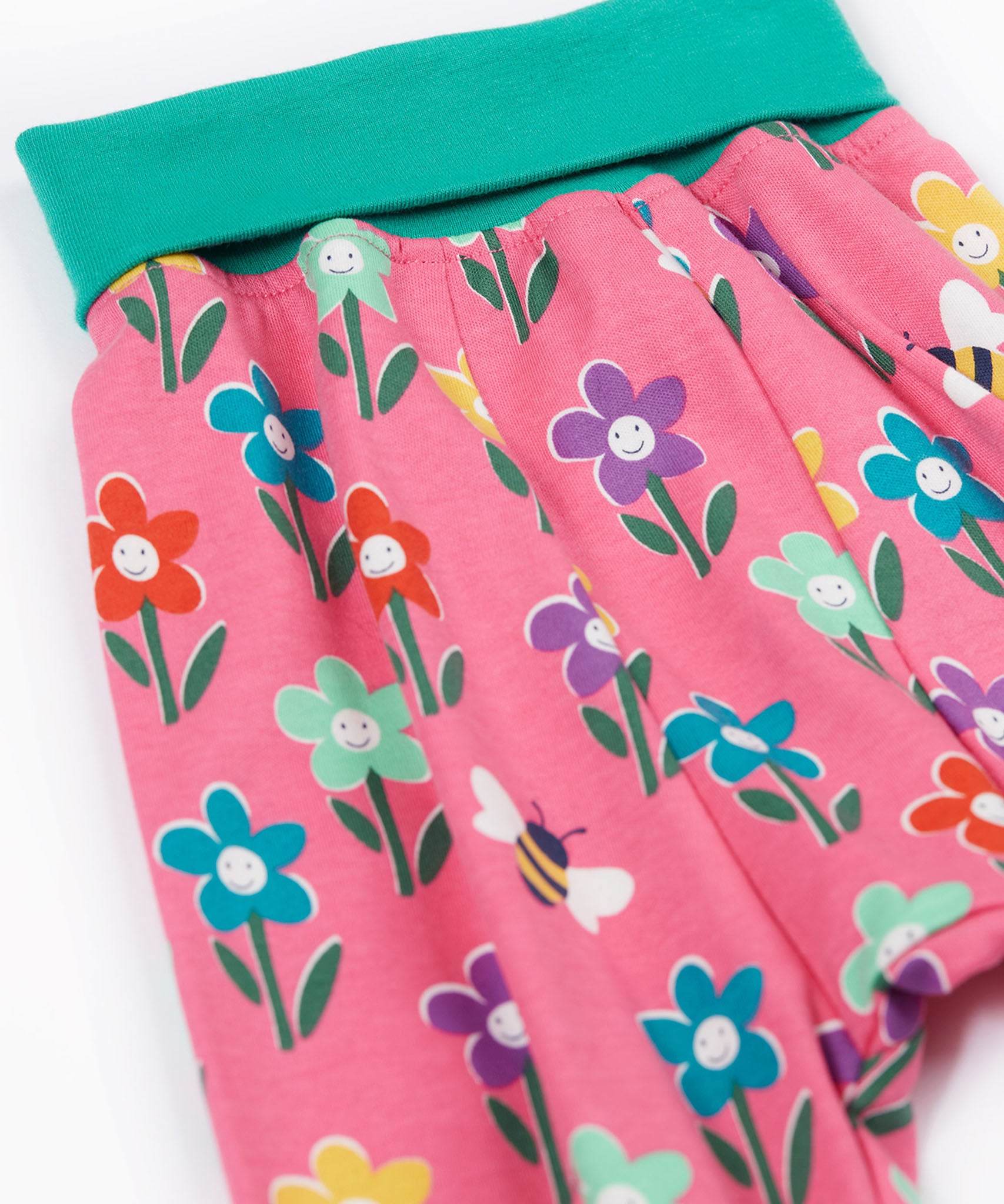 A close up of the print of the Frugi fun flowers parsnip pants. The fun flowers print features multi-coloured flowers and bees on a pink background with turquoise coloured waistband and leg cuffs.