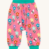 Frugi fun flowers print parsnip pants children's organic cotton trousers. The fun flowers print features multi-coloured flowers and bees on a pink background with turquoise coloured waistband and leg cuffs. 