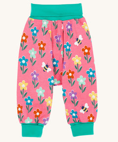 Frugi fun flowers print parsnip pants children's organic cotton trousers. The fun flowers print features multi-coloured flowers and bees on a pink background with turquoise coloured waistband and leg cuffs. 