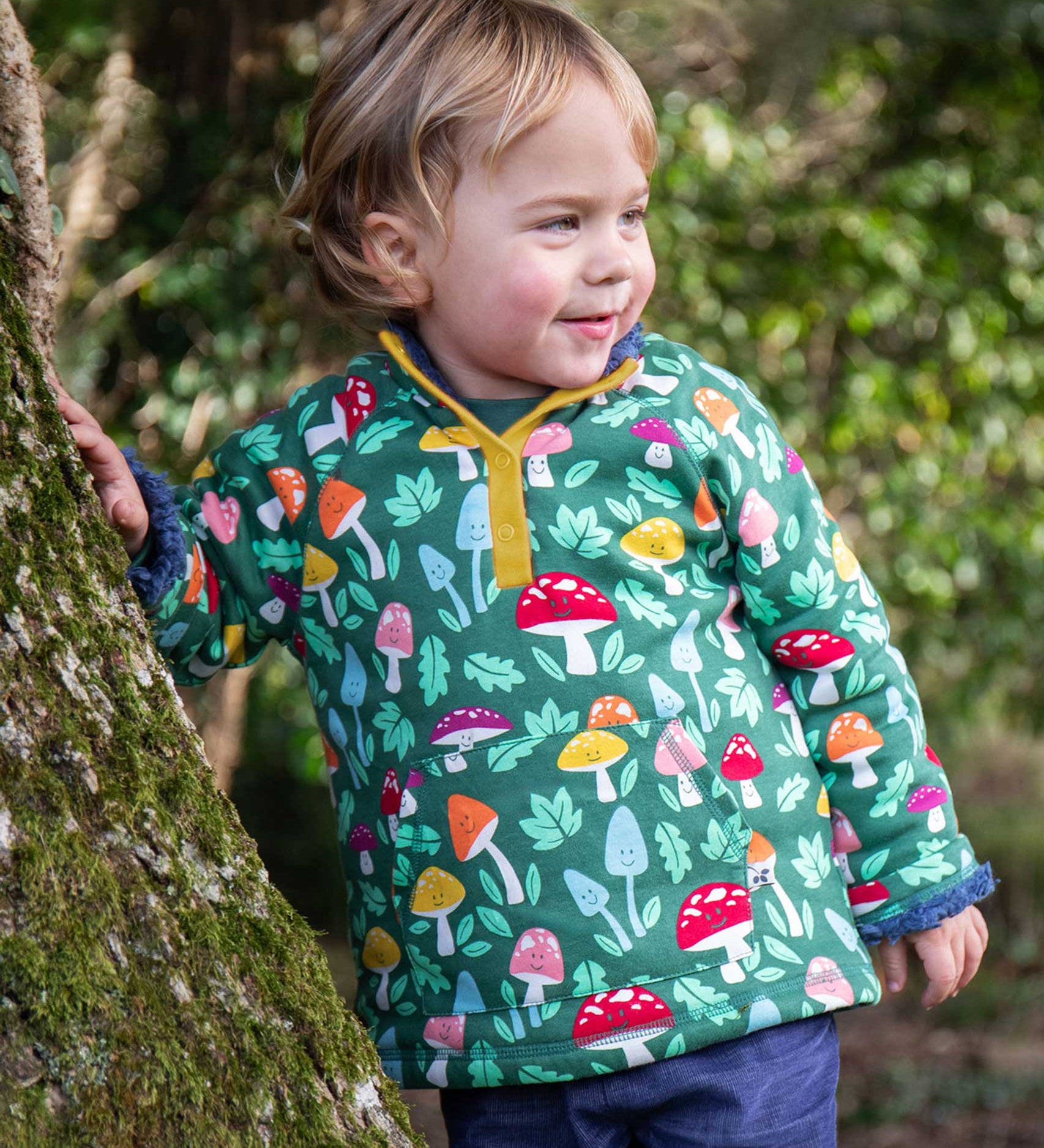 A child standing next to a tree wearing the Frugi Fun-Guys Children's Snuggle Fleece.