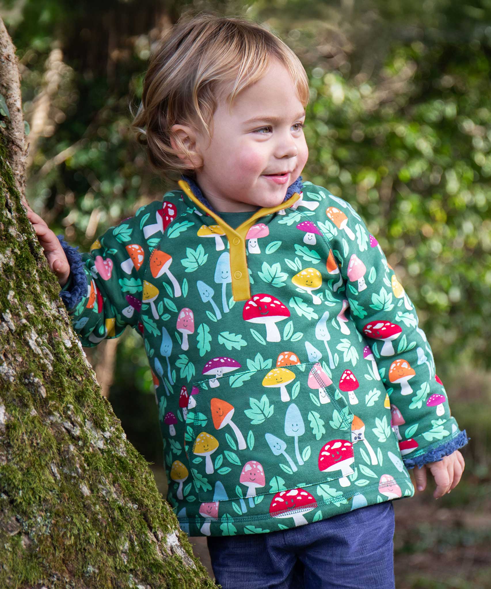 A child standing next to a tree wearing the Frugi Fun-Guys Children's Snuggle Fleece.