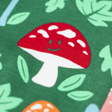 A close up of the red mushroom on the Frugi fun guys print dungarees.  The Fun guys print features multi-coloured mushrooms and light green leaves on a dark green background. The mushrroms all have happy faces.