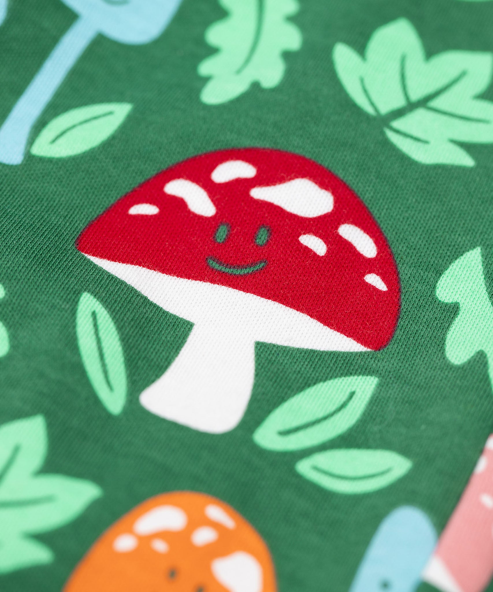 A close up of the red mushroom on the Frugi fun guys print dungarees.  The Fun guys print features multi-coloured mushrooms and light green leaves on a dark green background. The mushrroms all have happy faces.