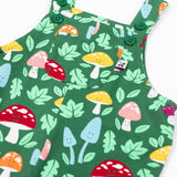 A close up of the front of the Frugi fun guys print dungarees with a pocket. The Fun guys print features multi-coloured mushrooms and light green leaves on a dark green background.