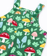 A close up of the front of the Frugi fun guys print dungarees with a pocket. The Fun guys print features multi-coloured mushrooms and light green leaves on a dark green background.