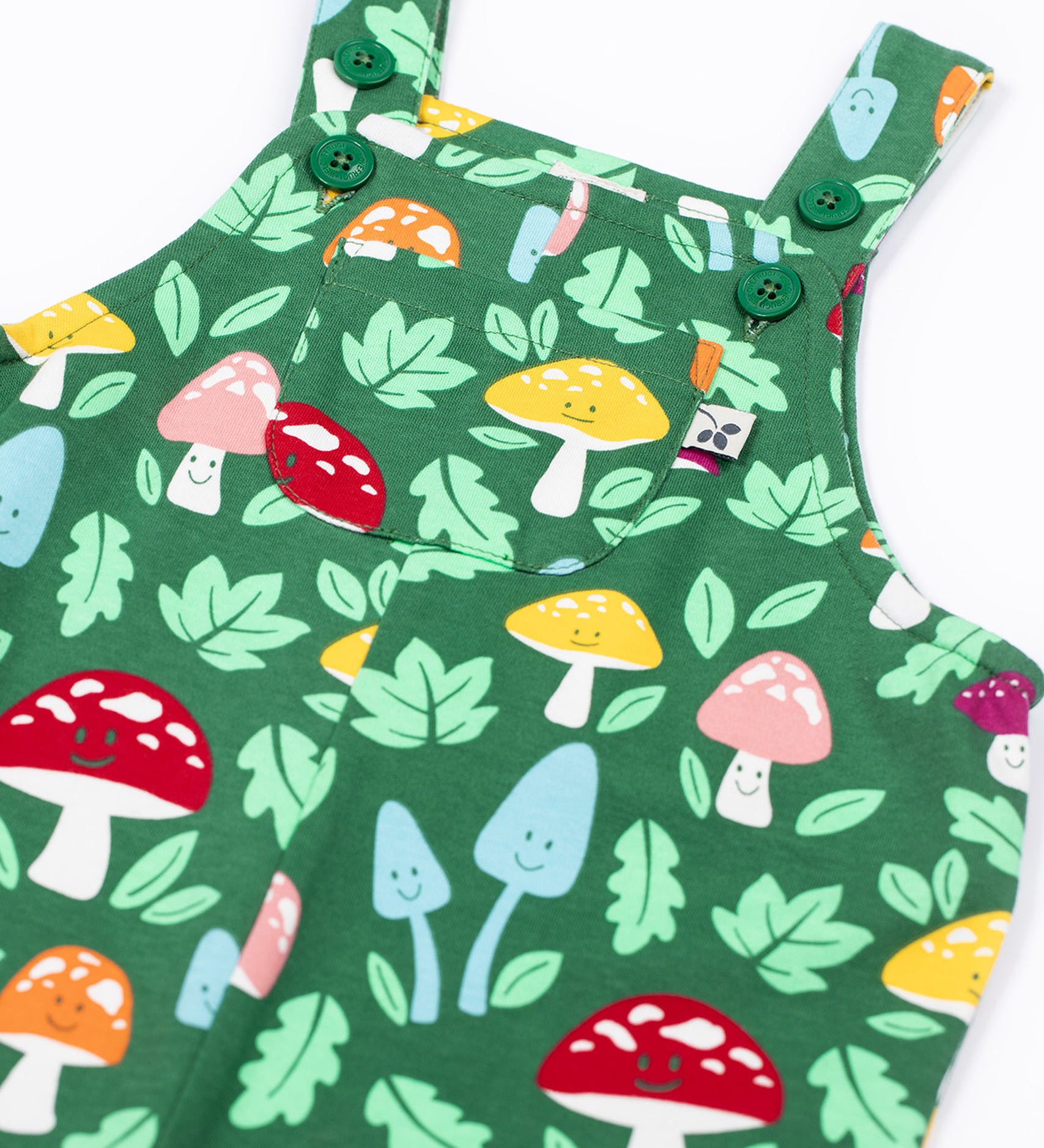 A close up of the front of the Frugi fun guys print dungarees with a pocket. The Fun guys print features multi-coloured mushrooms and light green leaves on a dark green background.