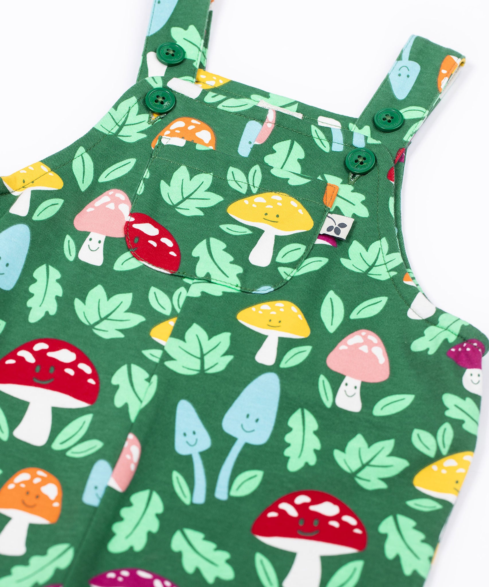 A close up of the front of the Frugi fun guys print dungarees with a pocket. The Fun guys print features multi-coloured mushrooms and light green leaves on a dark green background.