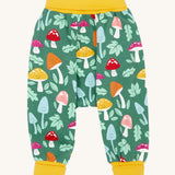 The Frugi fun guys multi coloured mushroom print parsnip pants children's trousers. The print features multi-coloured mushrooms, light green leaves on a dark green background with yellow cold coloured waistband and leg cuffs. 