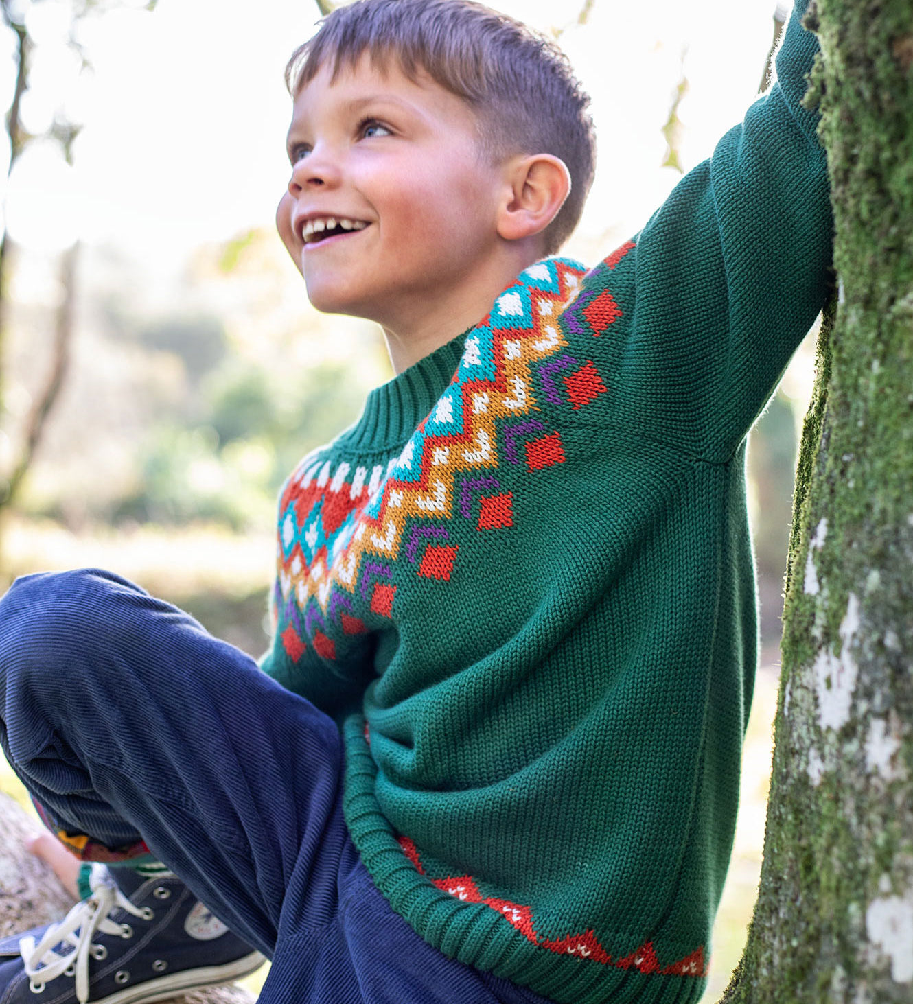 A child on a tree wearing the Frugi Fyfe Green Fair Isle Knitted Festive Jumper with navy cord trousers. The green kids jumper has a colourful Fair Isle pattern across the neck and chest and a border details on the bottom hem and sleeves. This Fair Isle jumper is a part of a wide range of organic cotton clothing for children available here at Babipur. 