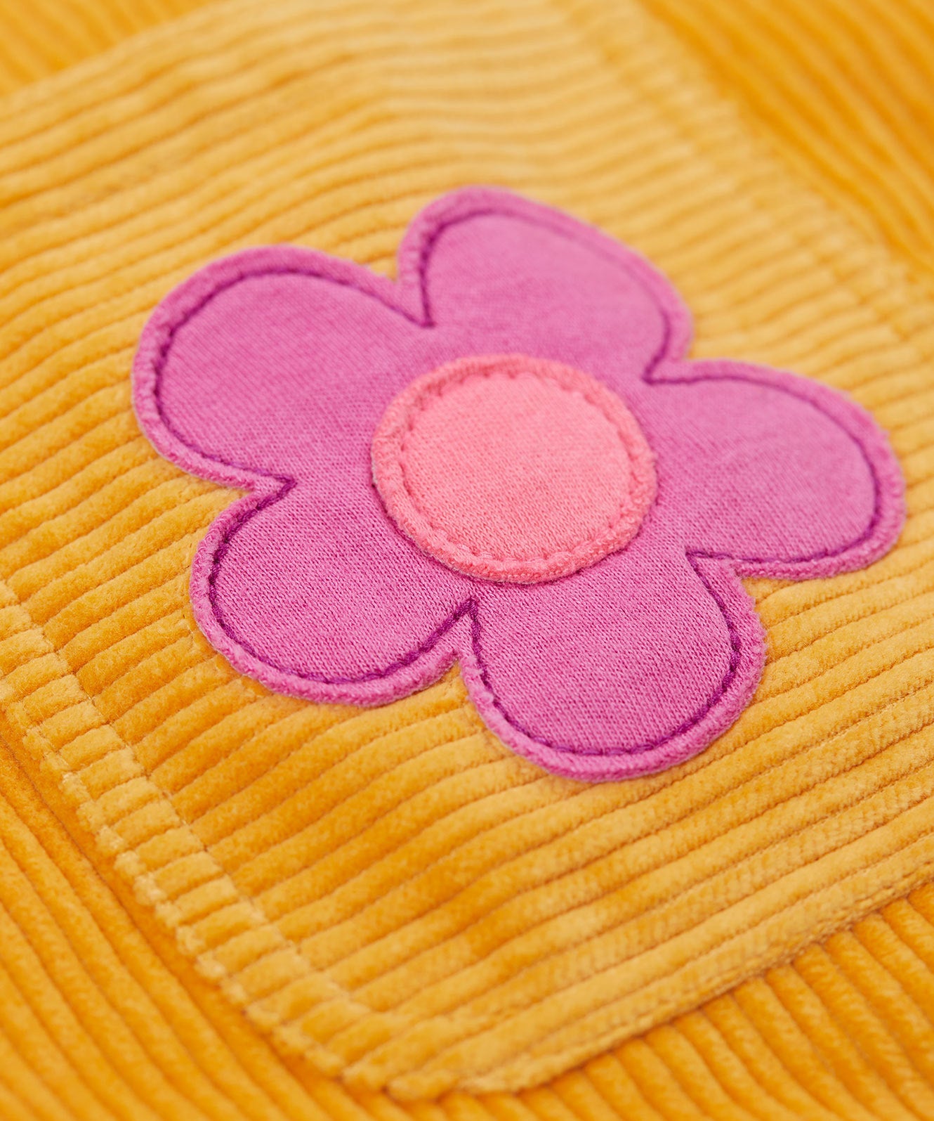 A close up of a pink flower on the Frugi organic cotton Ginny yellow gold Corduroy Pinafore Dress with Flowers with retro flower tights and a rainbow striped top underneath. This dress is a part of a wide range of organic cotton clothing for children available here at Babipur. 
