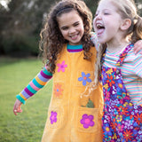 A child wearing the Frugi organic cotton Ginny yellow gold Corduroy Pinafore Dress with Flowers with retro  flower tights and a rainbow striped top underneath. This dress is a part of a wide range of organic cotton clothing for children available here at Babipur. 