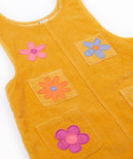 A close up of the Frugi organic cotton Ginny yellow gold Corduroy Pinafore Dress with Flowers with retro  flower tights and a rainbow striped top underneath. This dress is a part of a wide range of organic cotton clothing for children available here at Babipur. 