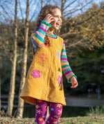 A child wearing the Frugi organic cotton Ginny yellow gold Corduroy Pinafore Dress with Flowers with retro  flower tights and a rainbow striped top underneath. This dress is a part of a wide range of organic cotton clothing for children available here at Babipur. 