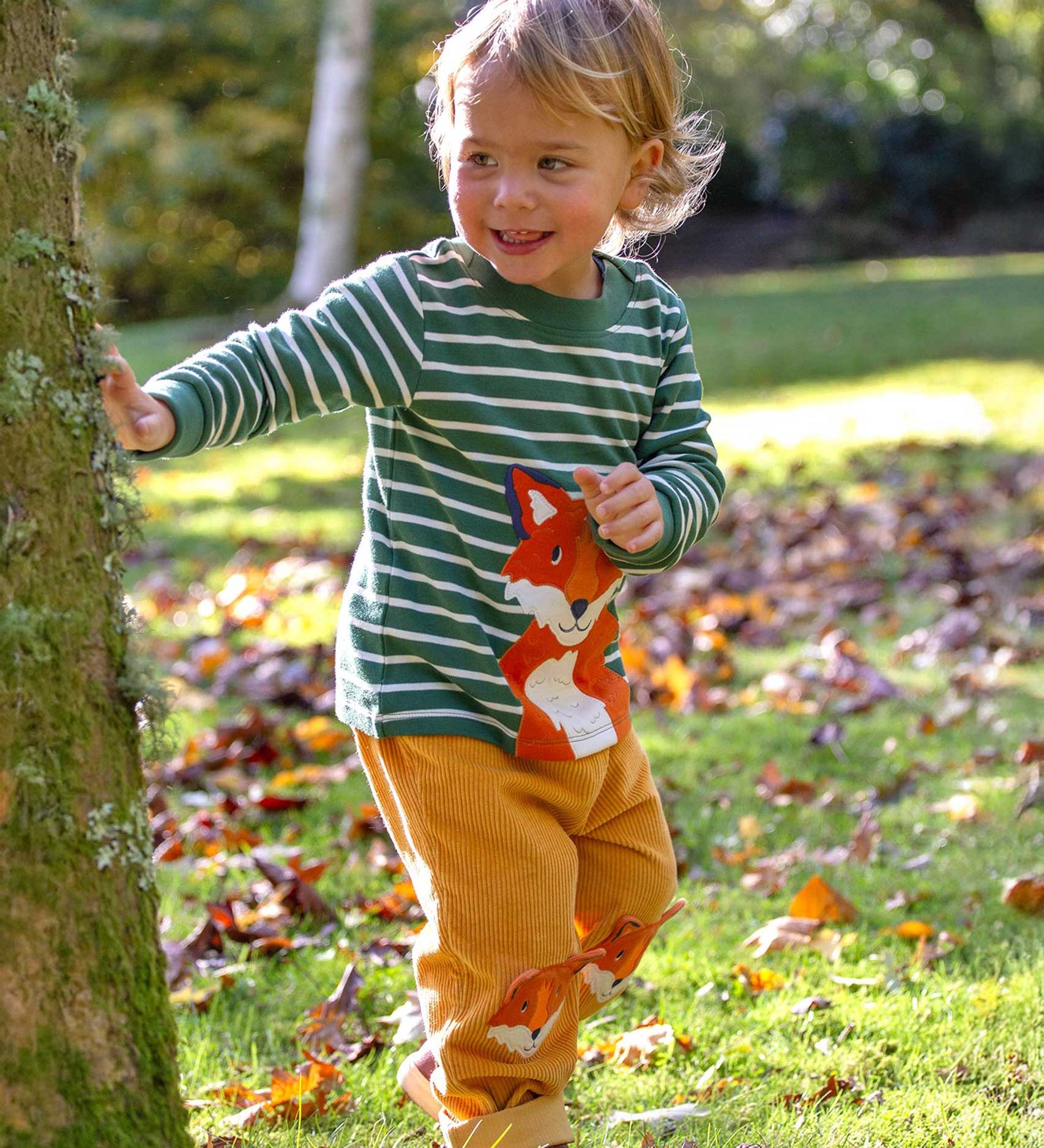 A child walking around outdoors wearing the Frugi easy on long sleeved top holly green breton stripe with fox applique and yellow coard fox trousers.