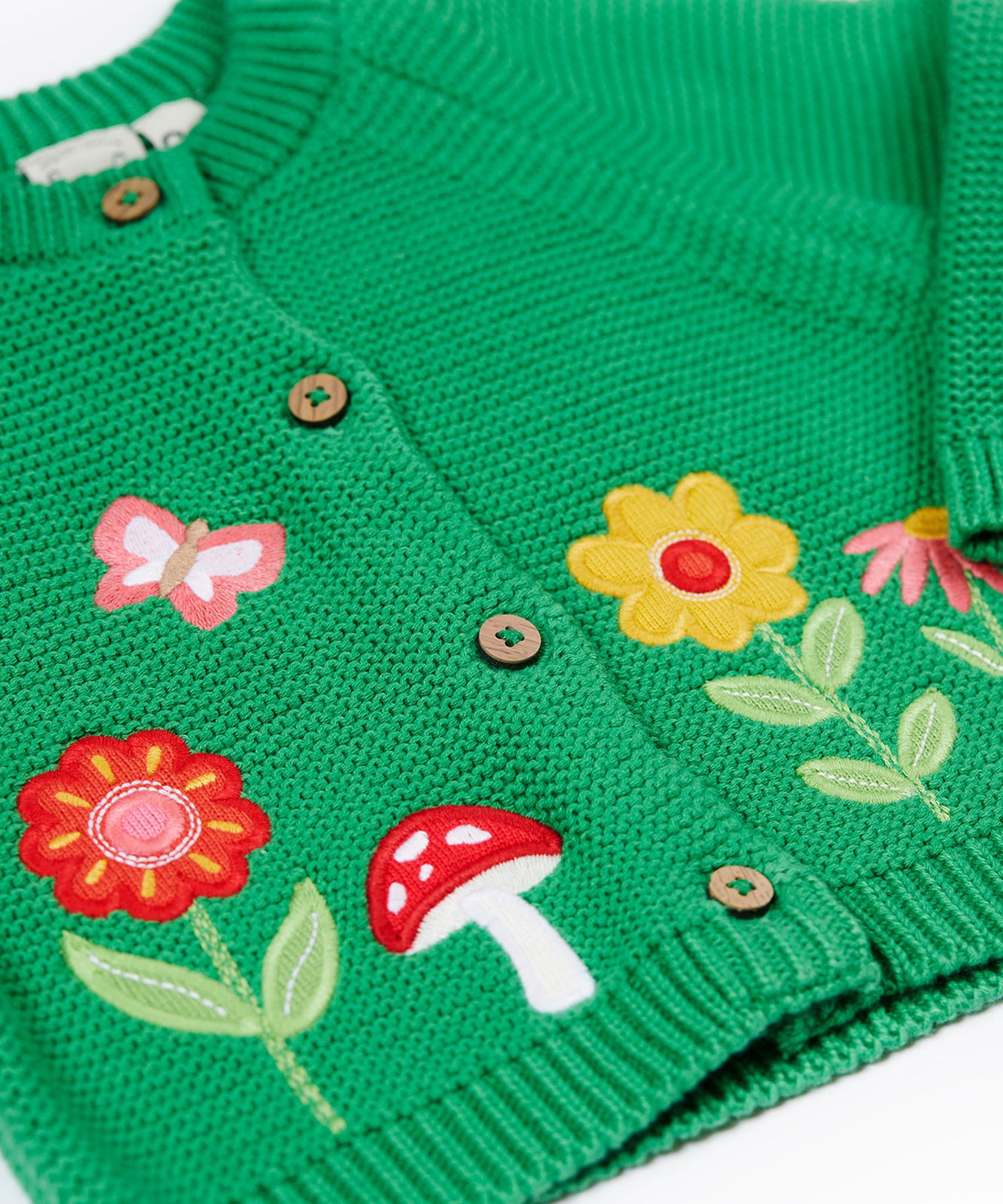 A close up of the flower, butterfly and mushroom appliques on the front of the Frugi colby clover green coloured knitted organic cotton cardigan.