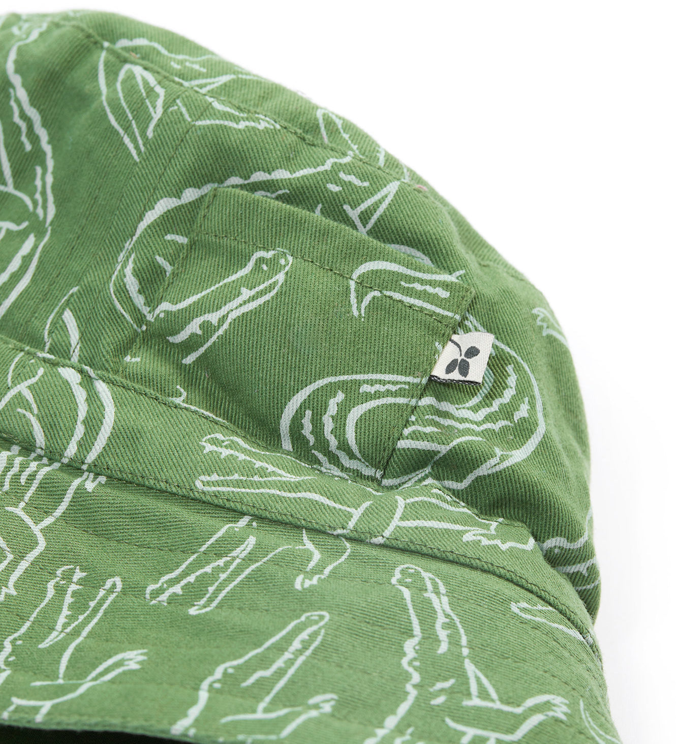 A close up of the Frugi kids green crocodile reversible hat. Showing the little pocket detail on the side of the hat surrounded by the white outlined crocodile design on green organic cotton fabric