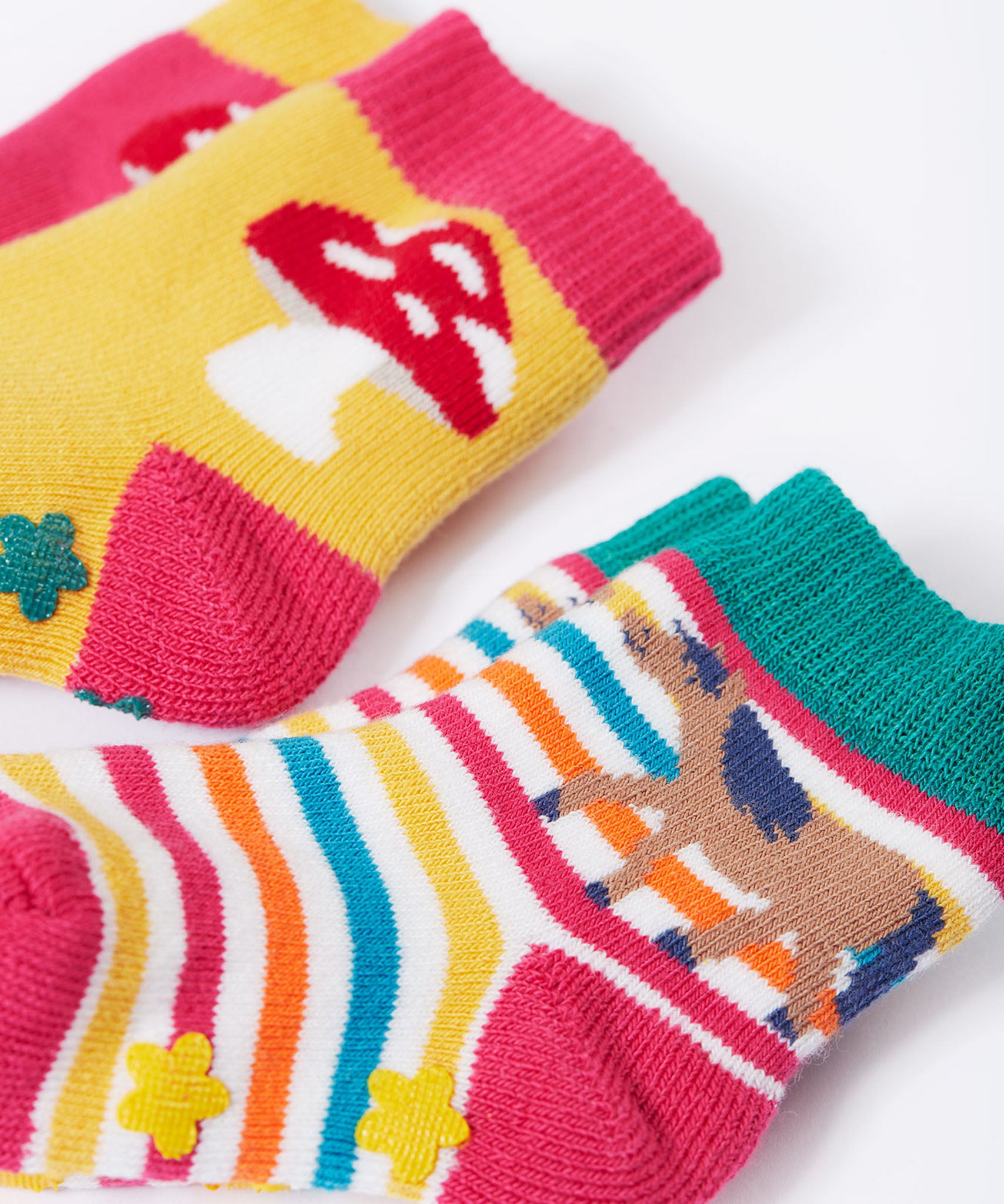 Frugi kids organic cotton grippy socks featuring wild woods designs.  One pair is rainbow striped with a horse and yellow flower grips. The other pair features a mushroom design with green flower grips, one sock is yellow one is pink.