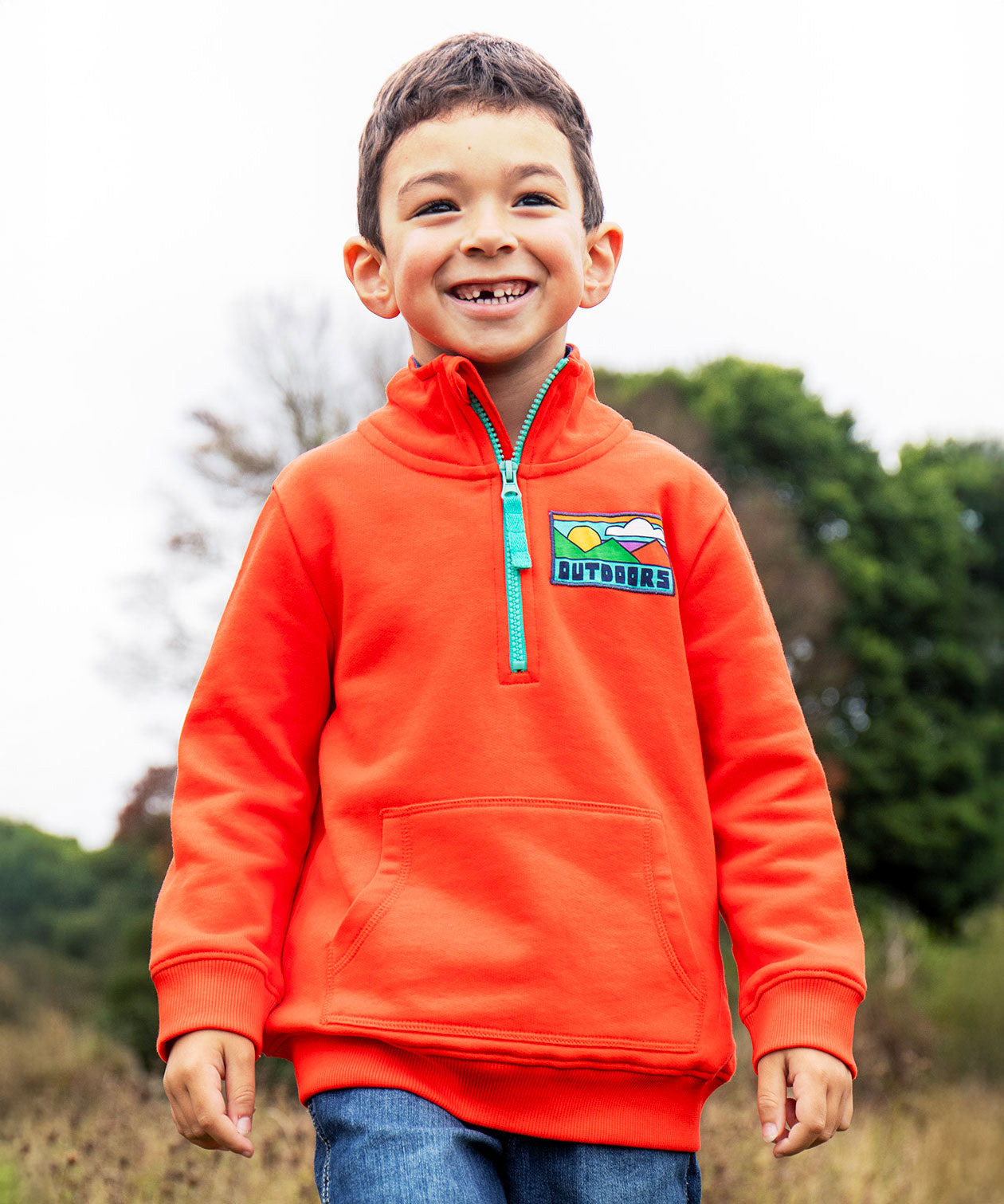A child wearing the Frugi Half Zip Organic Cotton Papaya orange Jumper with large Outdoors colour block style graphic on the chest.