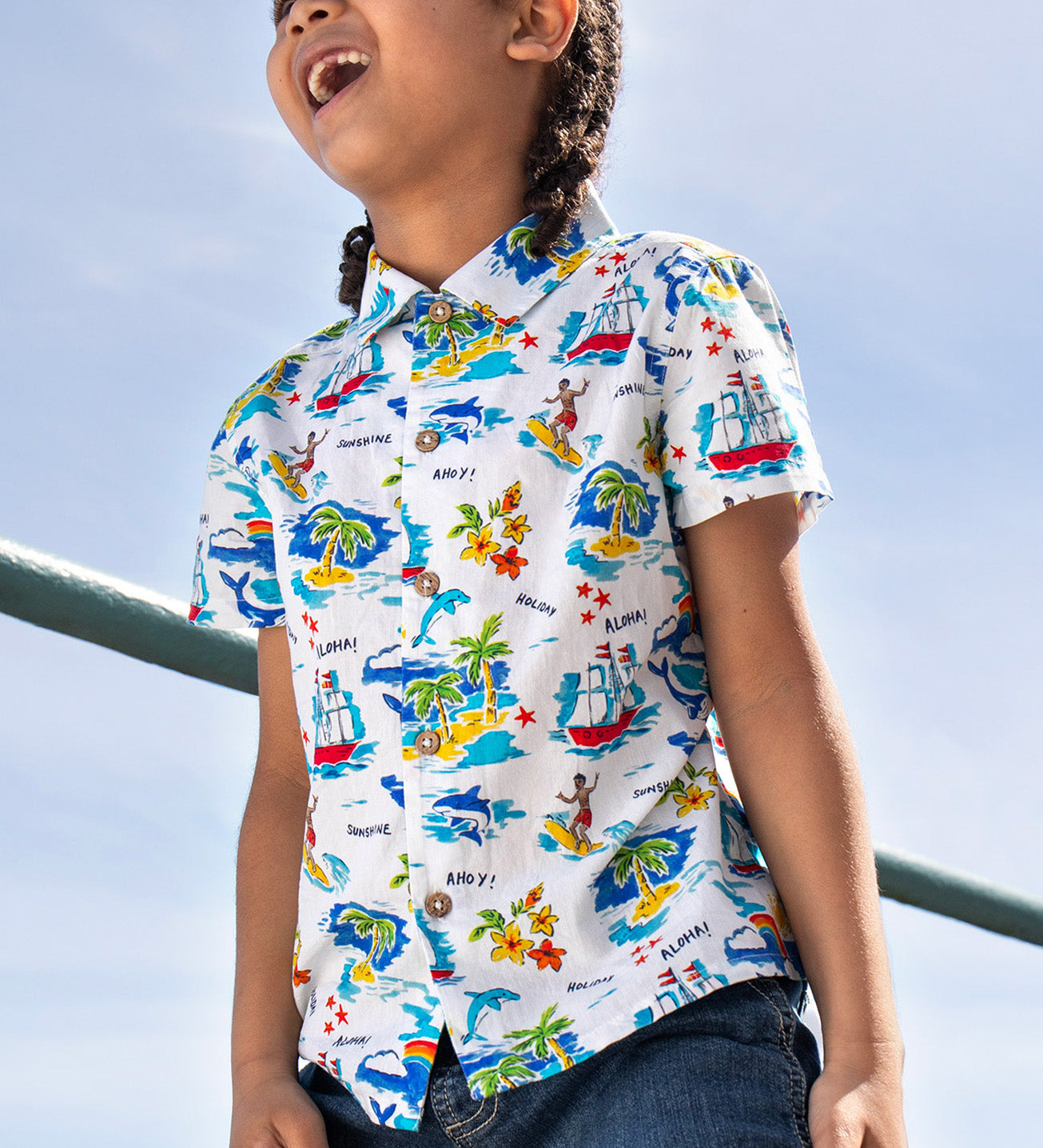 A laughing toddler wearing the Frugi Harvey Hawaiian Shirt in a Aloha print. Featuring a all-over tropical print with sailing ships, dolphins, rainbows, palm trees, surfers, and beach motifs on a white background, with wooden buttons down the middle, standing outside.
