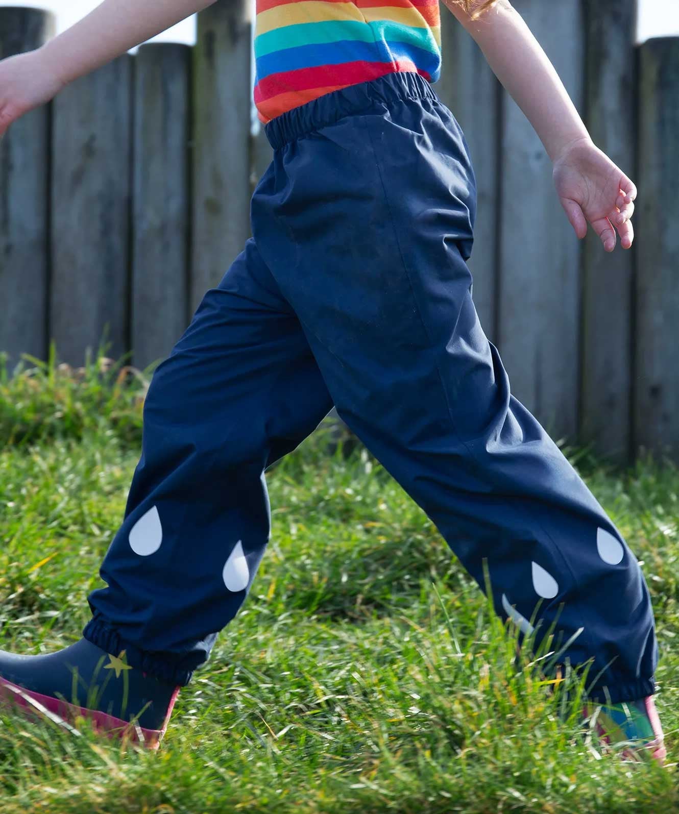 A close up of a child's legs, they are wearing the Frugi Indigo navy Rain Or Shine waterproof Trousers.The trousers have rain drop shaped reflective patches on the lower leg.