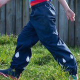 A close up of a child's legs, they are wearing the Frugi Indigo navy Rain Or Shine waterproof Trousers.The trousers have rain drop shaped reflective patches on the lower leg.