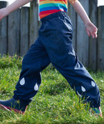 A close up of a child's legs, they are wearing the Frugi Indigo navy Rain Or Shine waterproof Trousers.The trousers have rain drop shaped reflective patches on the lower leg.