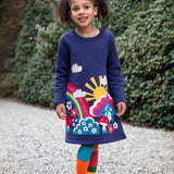 A child walking on a stone surface and wearing the Frugi kids kari dress in navy/rainbow.
