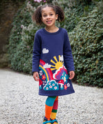 A child walking on a stone surface and wearing the Frugi kids kari dress in navy/rainbow.