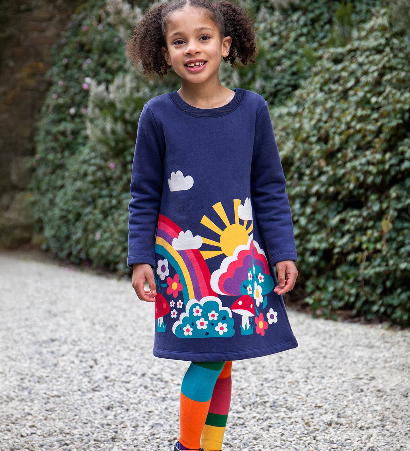 A child walking on a stone surface and wearing the Frugi kids kari dress in navy/rainbow.