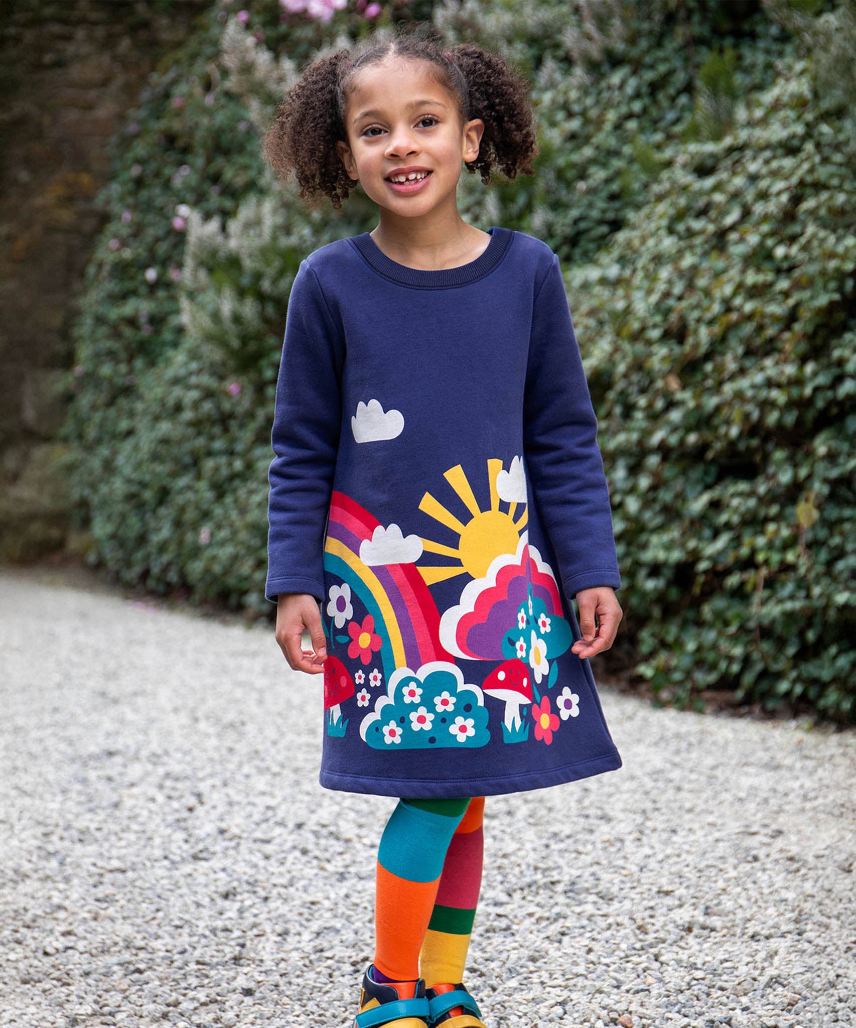 A child walking on a stone surface and wearing the Frugi kids kari dress in navy/rainbow.