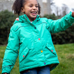 A child wearing the Frugi dragon/mushroom waterproof rambler coat. A thick and warm post consumer recycled polyester coat in bright turquoise with multicoloured popper fasteners. 