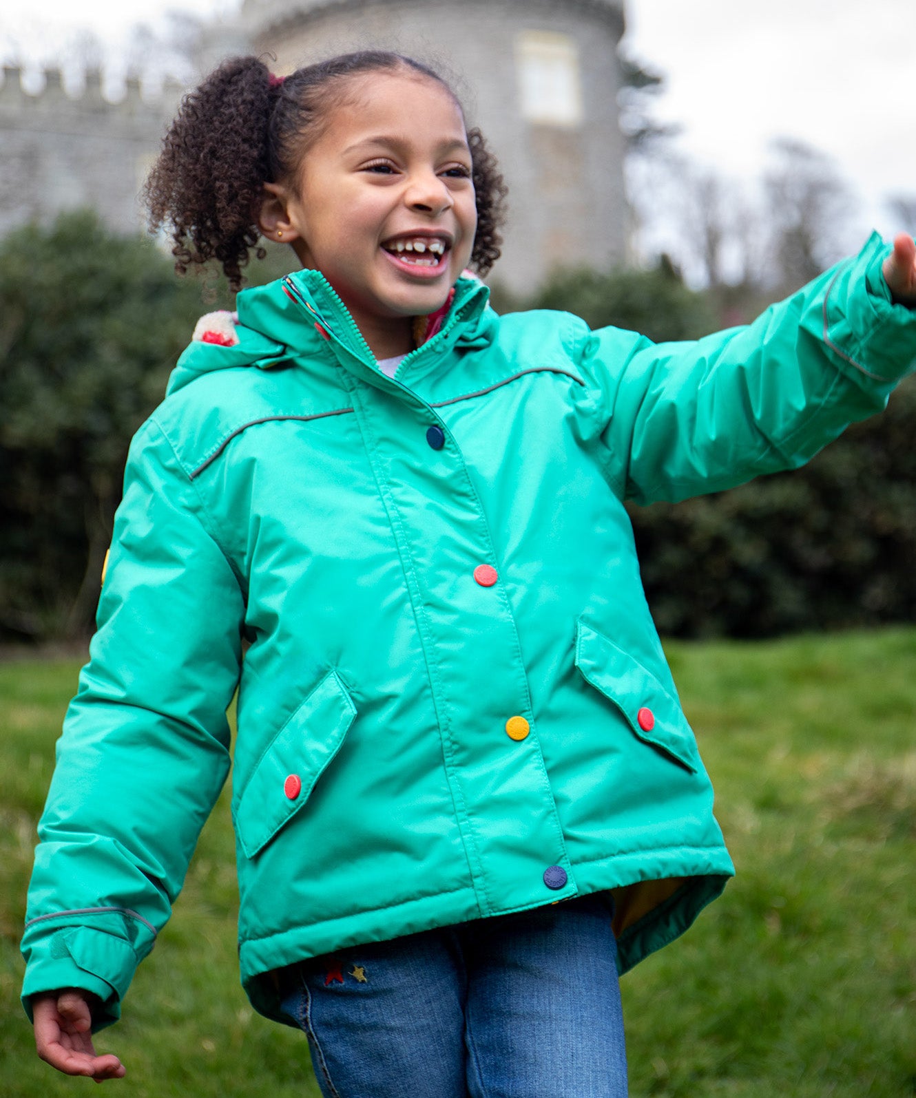 A child wearing the Frugi dragon/mushroom waterproof rambler coat. A thick and warm post consumer recycled polyester coat in bright turquoise with multicoloured popper fasteners. 