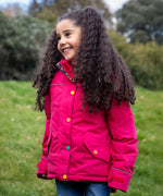 A child wearing the Frugi snapdragon/navy waterproof rambler coat. A post consumer recycled polyester coat in vibrant pink with multicoloured popper fasteners. 