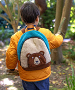 A child wearing the Frugi kids beary wild backpack outside showing scale. A blue and neutral colour fleece type with a corduroy character bear as a front pocket. Features a handy yellow carry handle on the top and brown corduroy shoulder straps