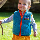 A toddler standing outside and wearing the Frugi bude borg fleece kids gilet in boat blue available at Babipur. A thick blue fleece with orange trim and a keep it wild slogan patch on the chest. 