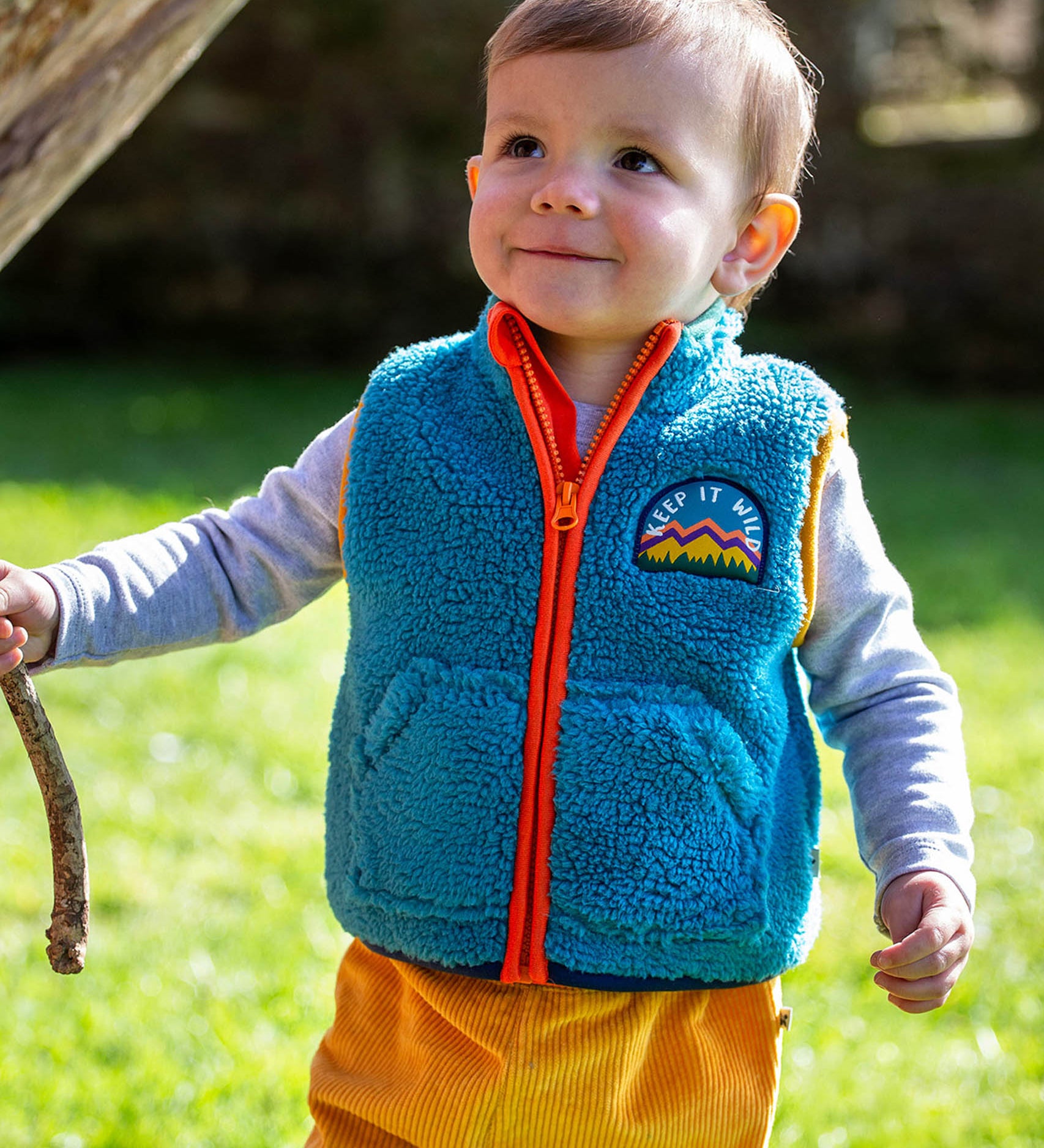 A toddler standing outside and wearing the Frugi bude borg fleece kids gilet in boat blue available at Babipur. A thick blue fleece with orange trim and a keep it wild slogan patch on the chest. 