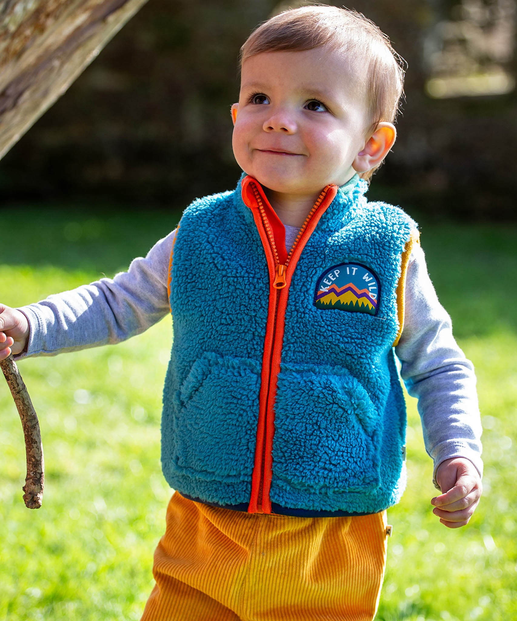 A toddler standing outside and wearing the Frugi bude borg fleece kids gilet in boat blue available at Babipur. A thick blue fleece with orange trim and a keep it wild slogan patch on the chest. 