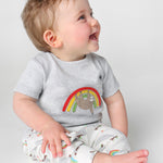 A happy toddler sat down and wearing the Frugi Children's GOTS Organic Cotton Frankie Summer Outfit - Sleepy Sloths. A beautiful 2 piece outfit including a grey short sleeved top with a sloth and rainbow character applique with comfy white cuffed sloth an