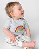 A happy toddler sat down and wearing the Frugi Children's GOTS Organic Cotton Frankie Summer Outfit - Sleepy Sloths. A beautiful 2 piece outfit including a grey short sleeved top with a sloth and rainbow character applique with comfy white cuffed sloth an
