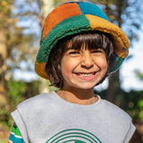 A child standing outside and wearing the Frugi kids fleece bucket hat in hotchpotch. A 100% recycled polyester thick fleeced bucket hat with a multicoloured block design